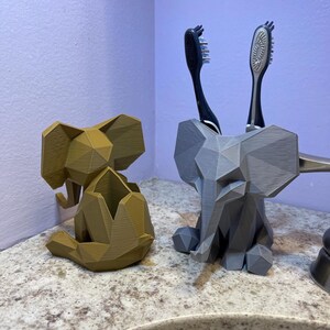 May include: Two geometric elephant-shaped bathroom organizers. One is a light brown color, and the other is gray. The gray elephant holds two toothbrushes. These decorative organizers add a unique touch to any bathroom.