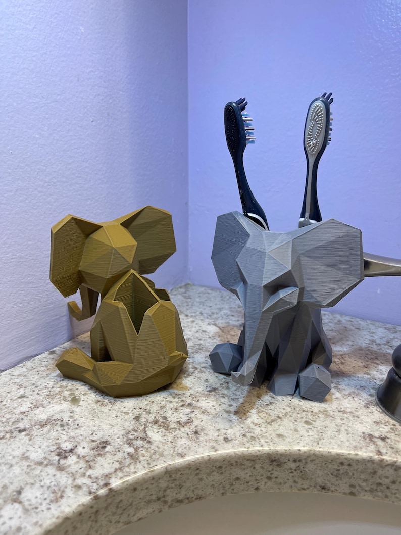 May include: Two geometric elephant-shaped bathroom organizers. One is a light brown color, and the other is gray. The gray elephant holds two toothbrushes. These decorative organizers add a unique touch to any bathroom.