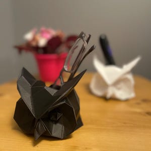 May include: A black and white owl-shaped desk organizer with a pair of glasses resting in the owl's beak. The owl is made of a 3D printed material and has a geometric design.