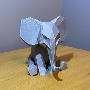 May include: A gray, geometric elephant figurine. The elephant is made of angular shapes, giving it a modern, low-poly design. It has large ears, a trunk, and is sitting down. The figurine is on a wooden surface.