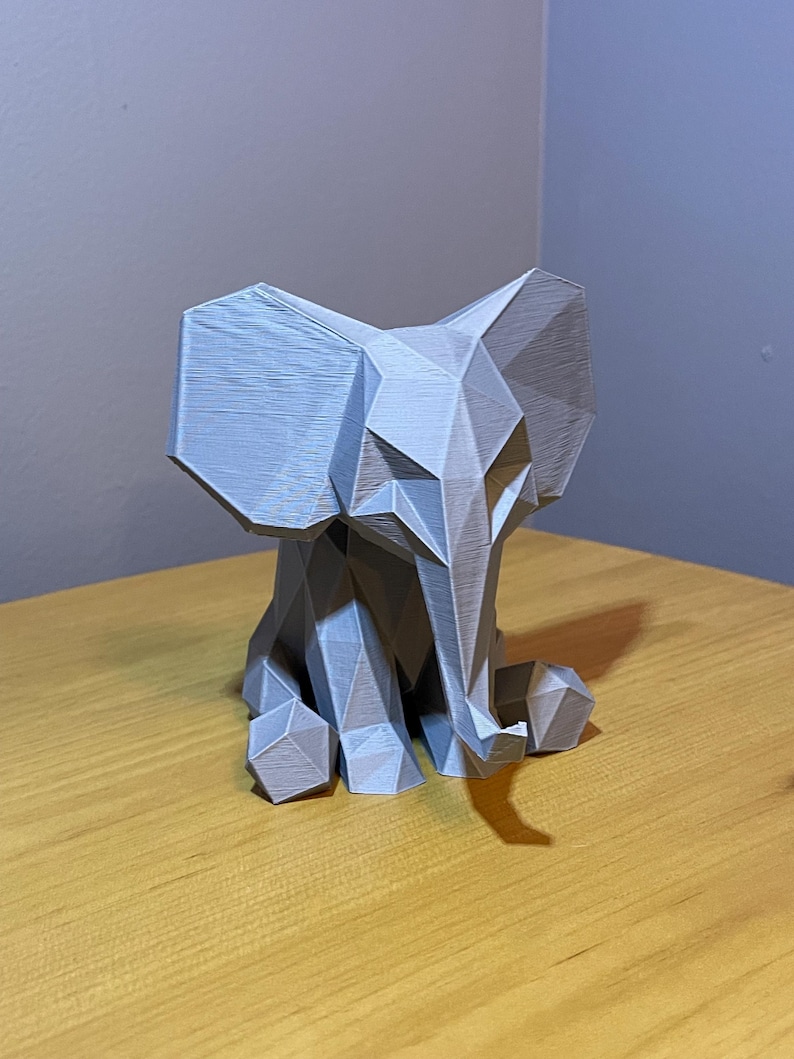 May include: A gray, geometric elephant figurine. The elephant is made of angular shapes, giving it a modern, low-poly design. It has large ears, a trunk, and is sitting down. The figurine is on a wooden surface.