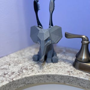 May include: A gray, geometric elephant toothbrush holder with two toothbrushes. The holder is made of a 3D-printed material and sits on a speckled countertop. The toothbrushes have black and silver handles and bristles.