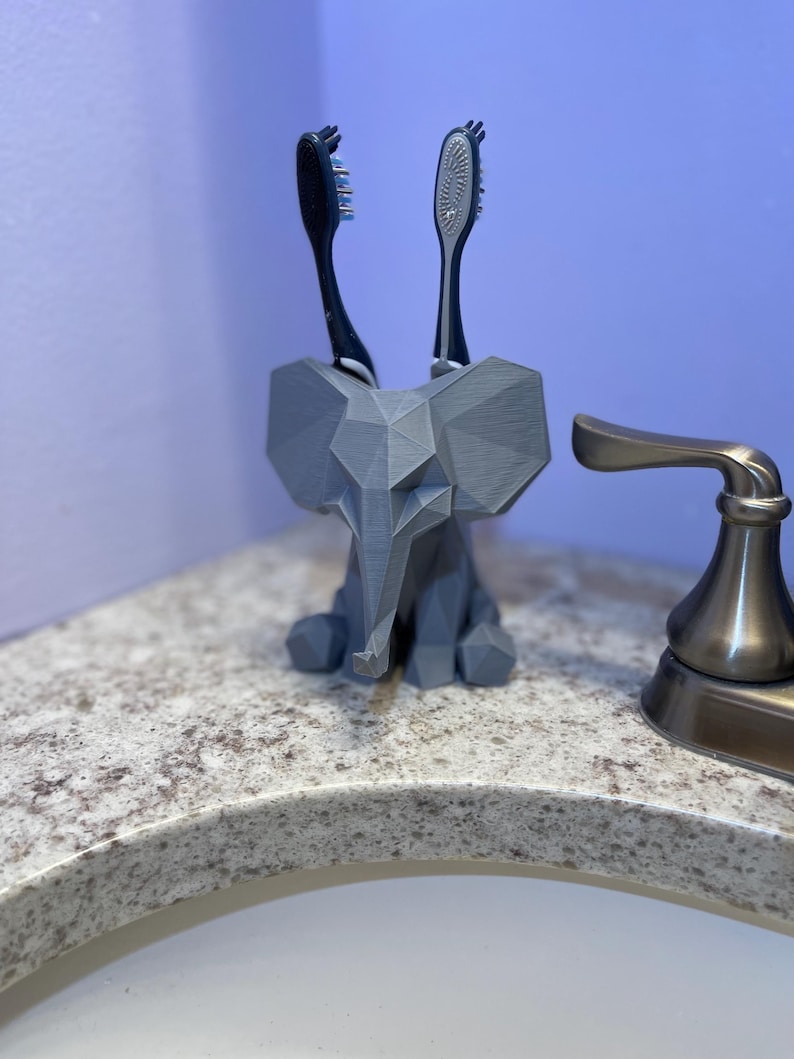 May include: A gray, geometric elephant toothbrush holder with two toothbrushes. The holder is made of a 3D-printed material and sits on a speckled countertop. The toothbrushes have black and silver handles and bristles.