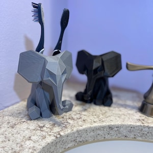 May include: A gray 3D-printed elephant toothbrush holder with two toothbrushes. A black elephant toothbrush holder is next to the gray one. The holders are on a speckled countertop near a sink.