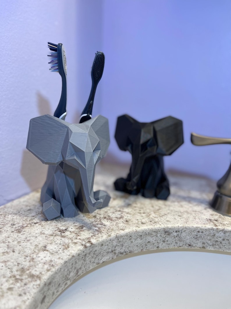 May include: A gray 3D-printed elephant toothbrush holder with two toothbrushes. A black elephant toothbrush holder is next to the gray one. The holders are on a speckled countertop near a sink.