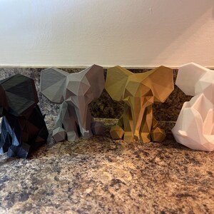 May include: Four geometric elephant figurines in black, gray, gold, and white. Each elephant is in a seated position with large ears and a detailed, angular design. The figurines are displayed on a speckled brown surface.