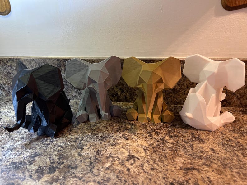 May include: Four geometric elephant figurines in black, gray, gold, and white. Each elephant is in a seated position with large ears and a detailed, angular design. The figurines are displayed on a speckled brown surface.