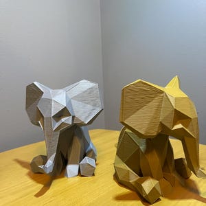 May include: Two geometric elephant figurines. One is light gray, the other is gold. Both are sitting with their trunks down and ears out. The figurines are made of a material with visible lines and angles, giving them a modern, polygonal look.