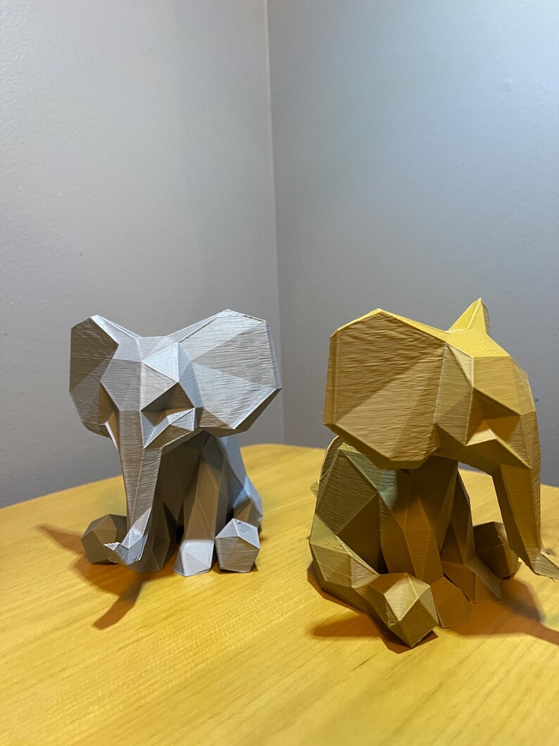 May include: Two geometric elephant figurines. One is light gray, the other is gold. Both are sitting with their trunks down and ears out. The figurines are made of a material with visible lines and angles, giving them a modern, polygonal look.