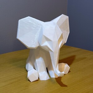 May include: A white, geometric elephant sculpture. The elephant is sitting with its trunk curled and ears spread. The sculpture has a faceted design, giving it a modern, minimalist aesthetic. The elephant is made of a solid white material.