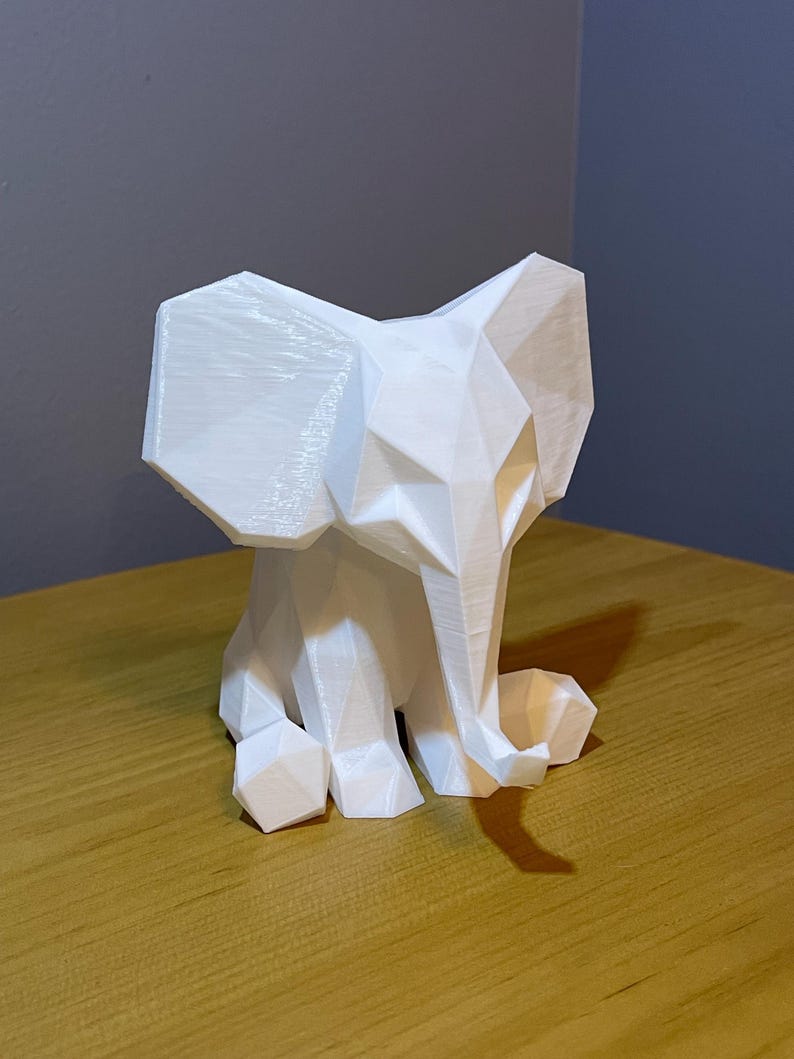 May include: A white, geometric elephant sculpture. The elephant is sitting with its trunk curled and ears spread. The sculpture has a faceted design, giving it a modern, minimalist aesthetic. The elephant is made of a solid white material.