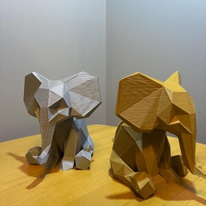 May include: Two geometric elephant figurines. One is light gray, the other is gold. Both are sitting on a wooden surface. The elephant figurines have a faceted design, giving them a modern, artistic look. The background is a neutral color.