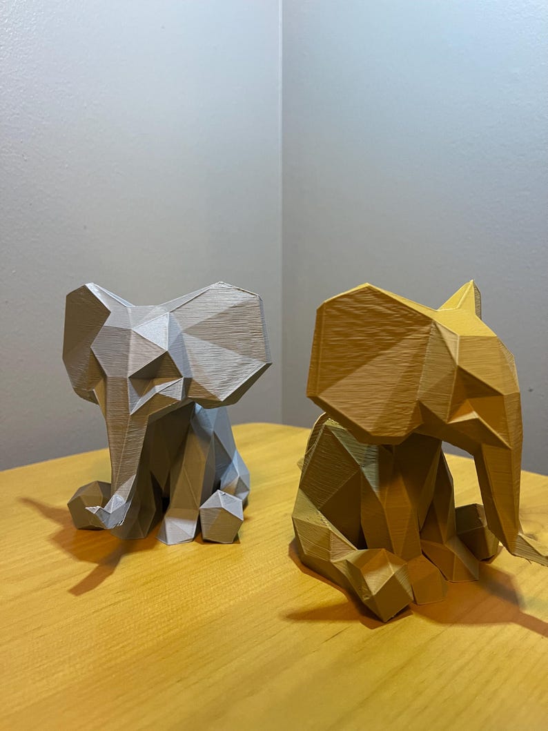 May include: Two geometric elephant figurines. One is light gray, the other is gold. Both are sitting on a wooden surface. The elephant figurines have a faceted design, giving them a modern, artistic look. The background is a neutral color.