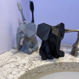 May include: Two geometric elephant toothbrush holders, one gray and one black, sit on a bathroom countertop. The gray holder has two toothbrushes, and the black holder is empty. The holders are made of a 3D-printed material.