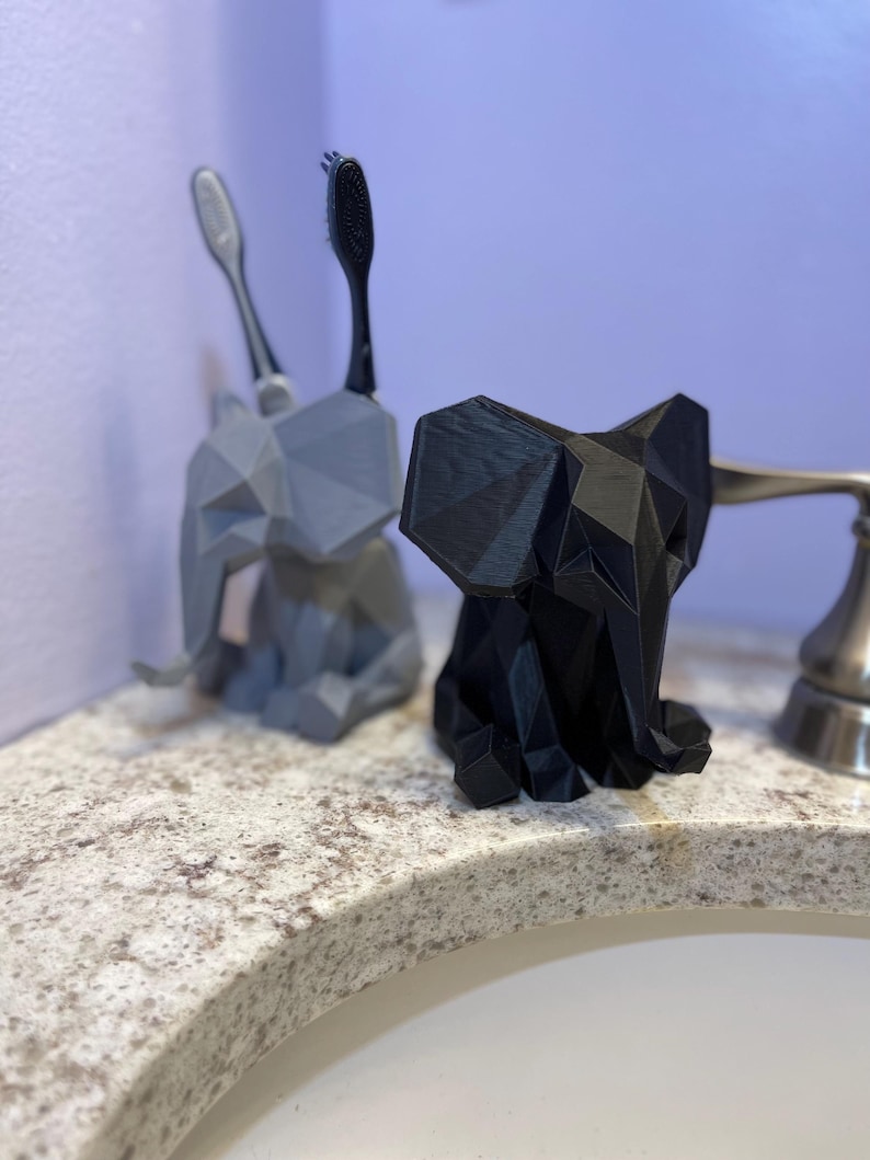 May include: Two geometric elephant toothbrush holders, one gray and one black, sit on a bathroom countertop. The gray holder has two toothbrushes, and the black holder is empty. The holders are made of a 3D-printed material.