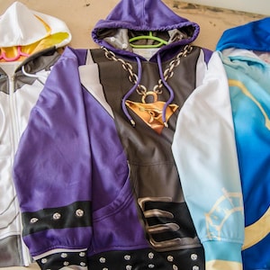 May include: Three zip-up hoodies in white, purple, and blue. The white hoodie has a geometric design, the purple one has a chain and pendant, and the blue one has a flowing pattern. All have hoods and are made of a soft material.