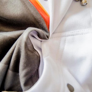 May include: Close-up of a white hoodie with a zipper and a pocket. The hoodie has a gray and black patterned design on the inside, with orange accents. The fabric is soft and appears to be a blend of materials.