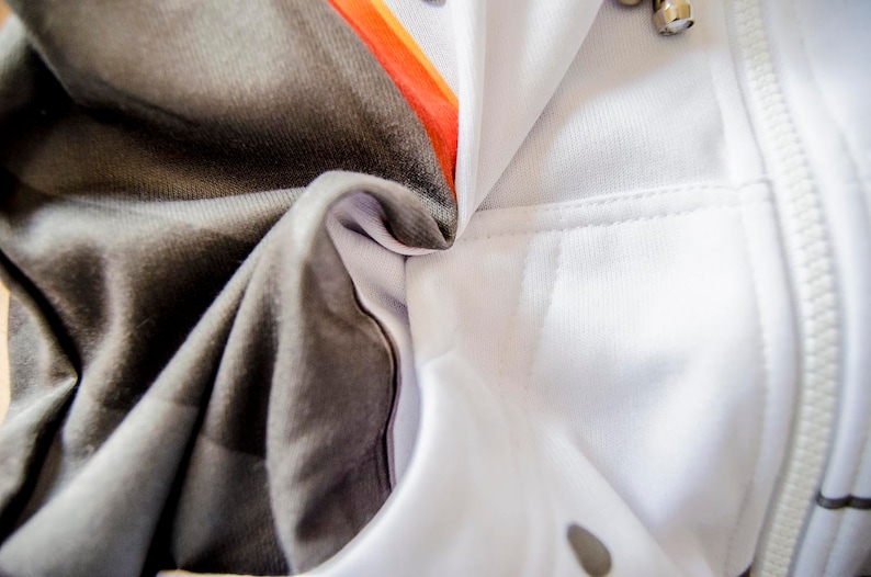 May include: Close-up of a white hoodie with a zipper and a pocket. The hoodie has a gray and black patterned design on the inside, with orange accents. The fabric is soft and appears to be a blend of materials.