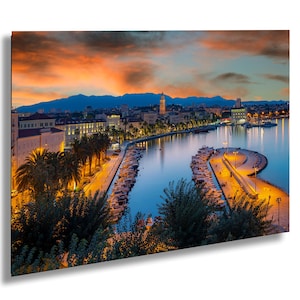 Może przedstawiać: Panoramiczny widok na panoramę miasta o zachodzie słońca z portem pełnym łodzi. Miasto jest oświetlone latarniami ulicznymi, a niebo jest w żywych pomarańczowych i różowych odcieniach.