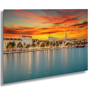 Peut inclure: Une vue panoramique d'un horizon urbain au coucher du soleil avec un front de mer et des palmiers. Le ciel est d'un orange et d'un rose vifs avec des nuages. Les bâtiments sont blancs et ont une architecture européenne classique. L'eau est calme et reflète les couleurs du ciel.