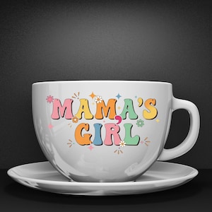 May include: A white ceramic coffee cup and saucer set. The cup features the text "MAMA'S GIRL" in colorful, retro-style lettering with floral and star accents. The cup has a large handle and sits on a matching saucer.