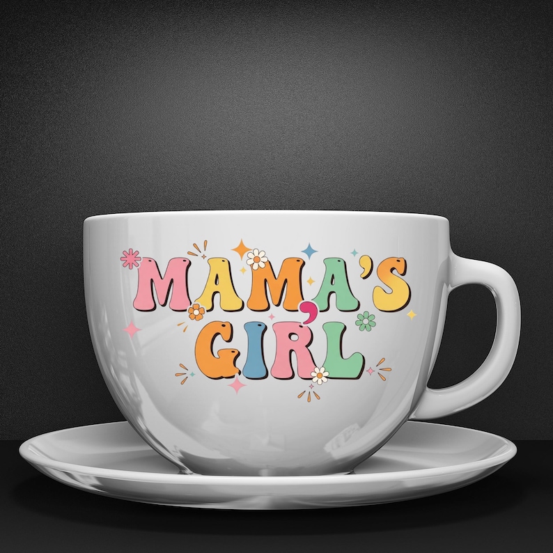 May include: A white ceramic coffee cup and saucer set. The cup features the text "MAMA'S GIRL" in colorful, retro-style lettering with floral and star accents. The cup has a large handle and sits on a matching saucer.