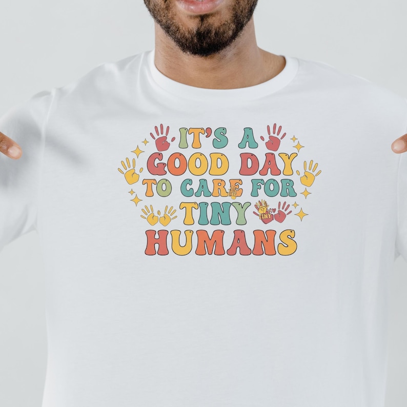 May include: White long-sleeve shirt with the text "IT'S A GOOD DAY TO CARE FOR TINY HUMANS" in a retro font. The design includes handprints and stars in various colors. A comfortable and stylish tee for caregivers.