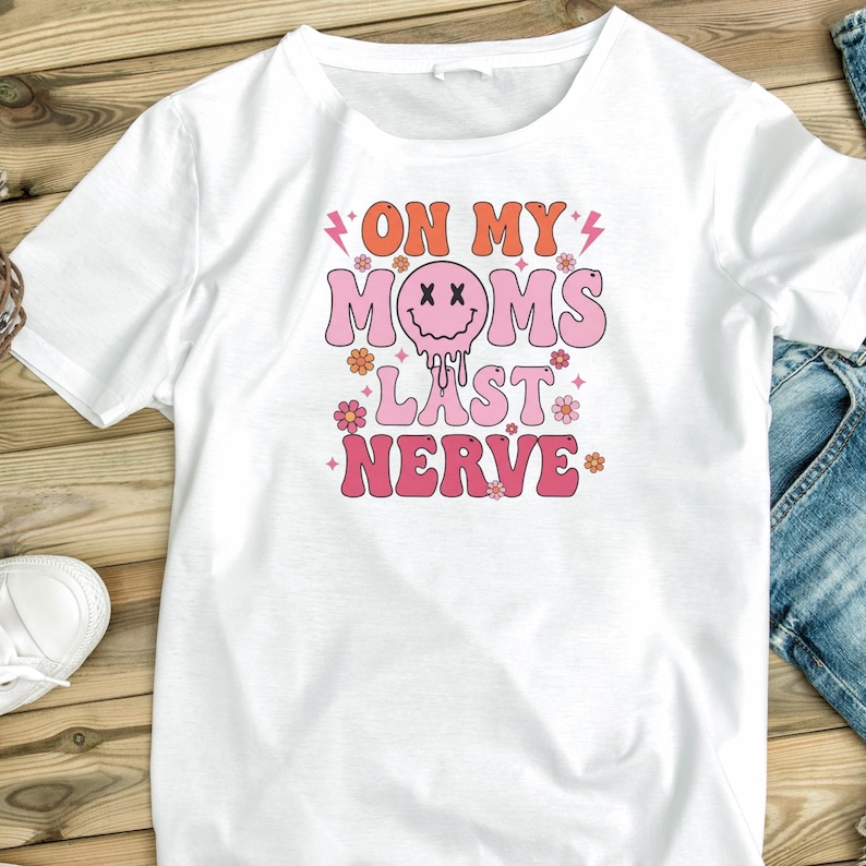 May include: White t-shirt with the text "ON MY MOMS LAST NERVE" in orange, pink, and white. The design includes a melting smiley face and floral accents. The shirt has short sleeves and a crew neck.