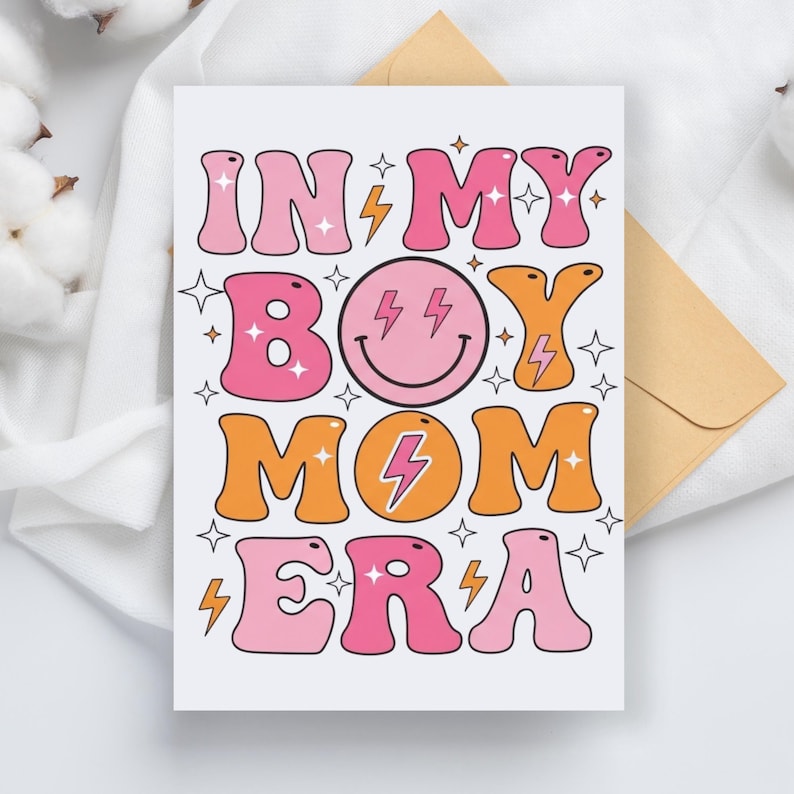 May include: A white greeting card with the text "IN MY BOY MOM ERA" in pink and orange bubble letters. The card features a smiley face with lightning bolts and star accents. An envelope is visible in the background.