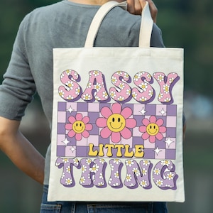 May include: A canvas tote bag with the words "SASSY LITTLE THING" in a retro font. The design includes pink flowers, a checkerboard pattern, and a smiley face. The bag has cream-colored handles.