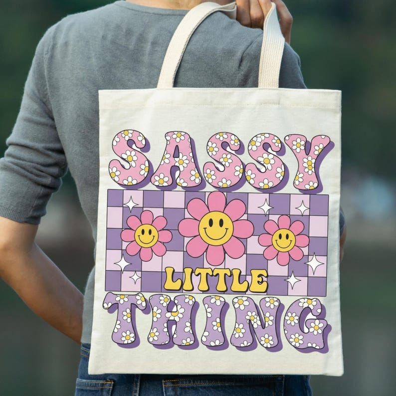 May include: A canvas tote bag with the words "SASSY LITTLE THING" in a retro font. The design includes pink flowers, a checkerboard pattern, and a smiley face. The bag has cream-colored handles.
