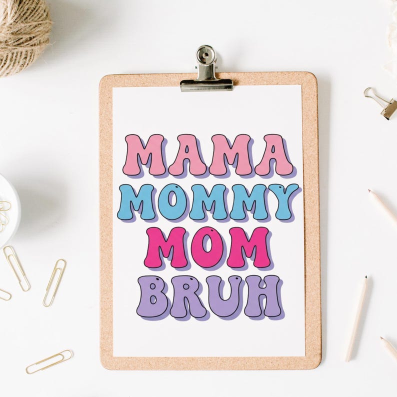 May include: A clipboard with a white paper featuring the words "MAMA MOMMY MOM BRUH" in pink, blue, and purple retro font. The clipboard is held by a silver clip. Other office supplies are scattered around the clipboard.