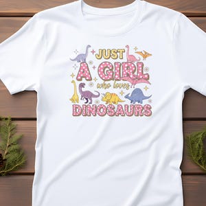 May include: White t-shirt featuring a colorful graphic design with the text "Just A Girl who loves Dinosaurs." The design includes various dinosaur illustrations in shades of pink, yellow, purple, and blue. The text is in a playful font with a polka dot pattern.