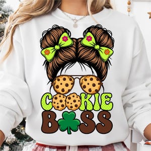 May include: White sweatshirt with a graphic design featuring a brown-haired character with green bows, cookie-shaped sunglasses, and the words "COOKIE BOSS" in green and brown. A green shamrock is also included in the design.