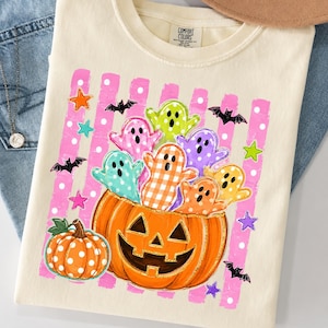 May include: A cream-colored t-shirt featuring a graphic of a large jack-o'-lantern filled with colorful ghosts in various patterns. The background includes pink vertical stripes with white polka dots, small bats, and stars. The label reads Comfort Colors.