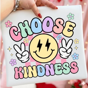 May include: A white square pillow with the words "CHOOSE KINDNESS" in colorful bubble letters. The design includes a yellow smiley face with lightning bolt eyes, peace signs, flowers, and a pink and white checkered background.