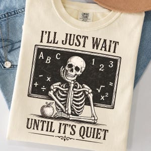 May include: A cream-coloured t-shirt featuring a black graphic of a skeleton sitting at a desk in front of a chalkboard. The text reads I'LL JUST WAIT UNTIL IT'S QUIET. The chalkboard displays letters, numbers, and math symbols, with an apple on the desk.