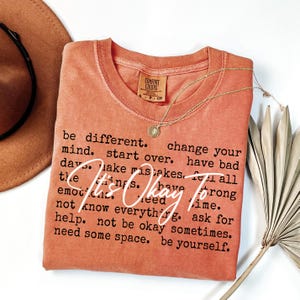 May include: A terracotta-colored short-sleeve t-shirt with a crew neckline. The shirt features black printed text that reads: be different. change your mind. start over. have bad days. make mistakes. feel all the things. have strong emotions. need time. not know everything. ask for help. not be okay sometimes. need some space. be yourself. Overlaid in white script is the text: It's Okay To.