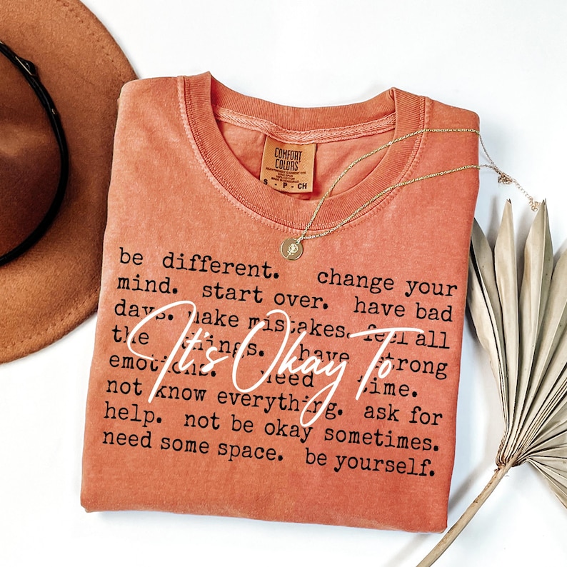 May include: A terracotta-colored short-sleeve t-shirt with a crew neckline. The shirt features black printed text that reads: be different. change your mind. start over. have bad days. make mistakes. feel all the things. have strong emotions. need time. not know everything. ask for help. not be okay sometimes. need some space. be yourself. Overlaid in white script is the text: It's Okay To.