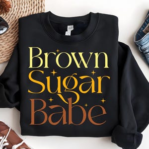 May include: Black sweatshirt with the text "Brown Sugar Babe" in yellow and orange lettering. The text is stylized with a vintage font and includes small star accents.