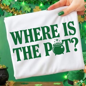 May include: White t-shirt with the green text "WHERE IS THE POT?" and a green pot of gold graphic. The shirt is held up by a hand with green nail polish. The background includes shamrock decorations and a pot of gold.