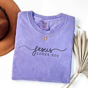 May include: A light purple short-sleeve t-shirt with a crew neckline. The shirt features the black text "jesus LOVES YOU" in a cursive and sans-serif font combination. A gold-toned necklace with a small circular pendant rests on the fabric.