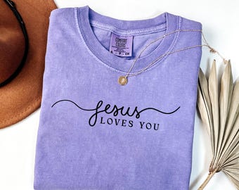 May include: A light purple short-sleeve t-shirt with a crew neckline. The shirt features the black text "jesus LOVES YOU" in a cursive and sans-serif font combination. A gold-toned necklace with a small circular pendant rests on the fabric.