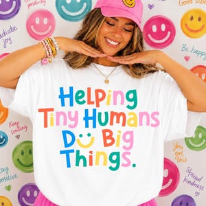 May include: A white t-shirt featuring the multicolored text Helping Tiny Humans Do Big Things, with a yellow smiley face replacing the letter O in Do. The shirt is paired with a pink baseball cap with a yellow smiley face on the front.