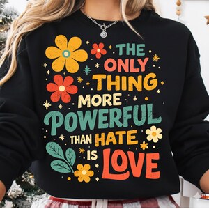 Può includere: Felpa nera con un design floreale e la scritta "THE ONLY THING MORE POWERFUL THAN HATE IS LOVE". Il design include fiori e stelle gialle, arancioni e turchesi.