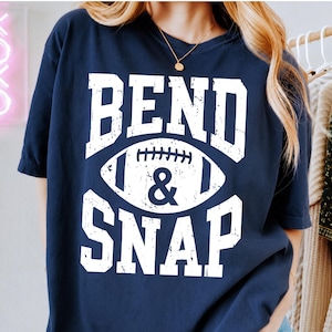 May include: A navy blue short-sleeved t-shirt featuring a distressed white graphic print. The design includes the text BEND & SNAP in a bold, block font, with a football illustration positioned between the two lines of text on the front.