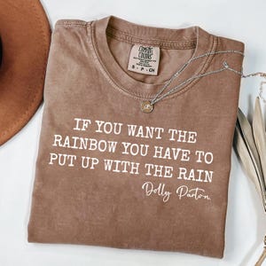 Könnte beinhalten: Ein braunes kurzärmeliges T-Shirt mit Rundhalsausschnitt und dem weißen Aufdruck: IF YOU WANT THE RAINBOW YOU HAVE TO PUT UP WITH THE RAIN Dolly Parton. Eine silberfarbene Halskette mit einem kleinen runden Anhänger liegt auf dem T-Shirt.