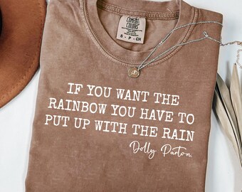 May include: A brown short-sleeved t-shirt with a crew neckline featuring the white printed text: IF YOU WANT THE RAINBOW YOU HAVE TO PUT UP WITH THE RAIN Dolly Parton. A silver-toned chain necklace with a small circular pendant rests on the shirt.