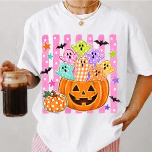 May include: A white t-shirt featuring a graphic of a large orange jack-o'-lantern filled with colorful ghosts in various patterns, surrounded by small bats and stars against a background of vertical pink stripes with white polka dots.