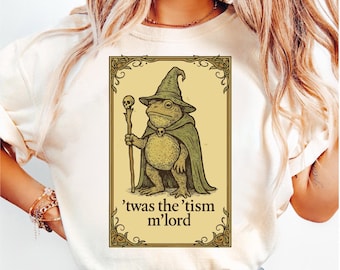 May include: Cream-colored t-shirt featuring a whimsical illustration of a frog dressed as a wizard, holding a staff with a skull. The frog wears a pointed hat and cape. The text "'twas the 'tism m'lord" is below the illustration.